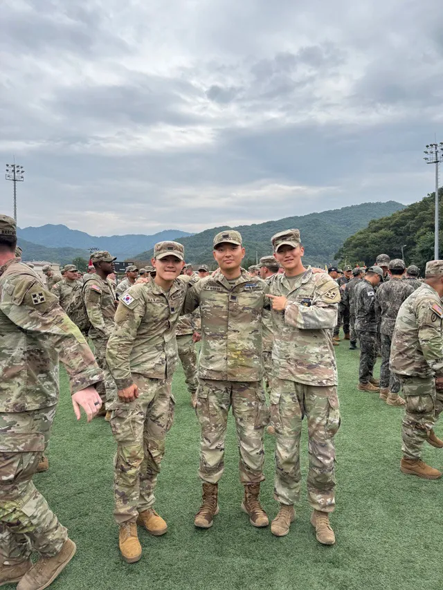 Doheon Kim standing with two fellow soldiers on the field after the badge ceremony, mountains behind them