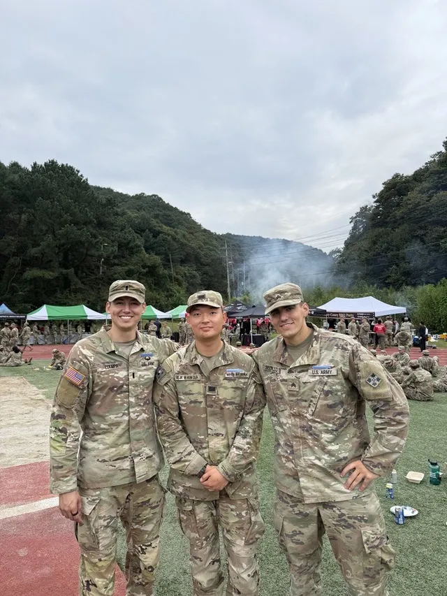 Doheon Kim between two fellow soldiers in front of event tents, with wooded hills behind