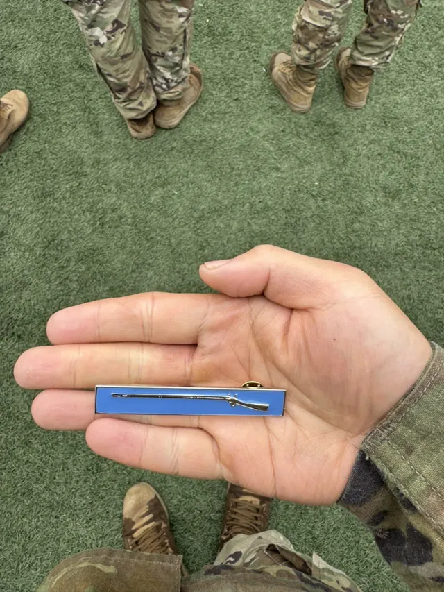 An Expert Infantryman Badge, a silver rifle on a blue bar, resting in an open palm above the turf