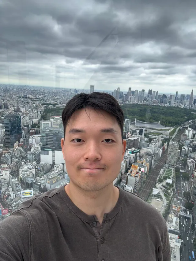 Doheon Kim taking a selfie behind glass at Shibuya Sky, with a park and a skyline of towers behind him under grey clouds