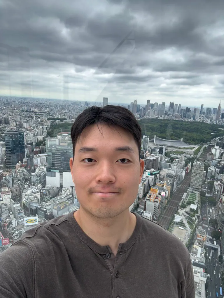 Doheon Kim taking a selfie behind glass at Shibuya Sky, with a park and a skyline of towers behind him under grey clouds