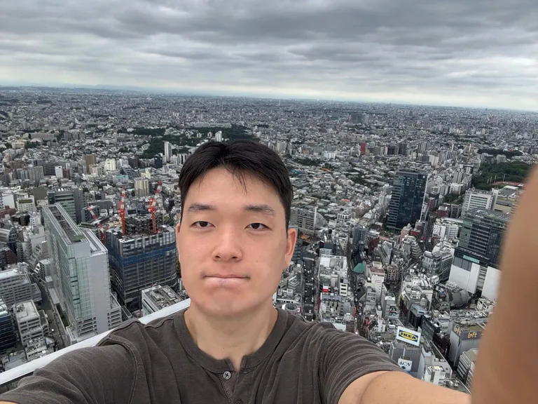 Doheon Kim taking a selfie on the Shibuya Sky rooftop, with dense city blocks stretching to the horizon under grey clouds