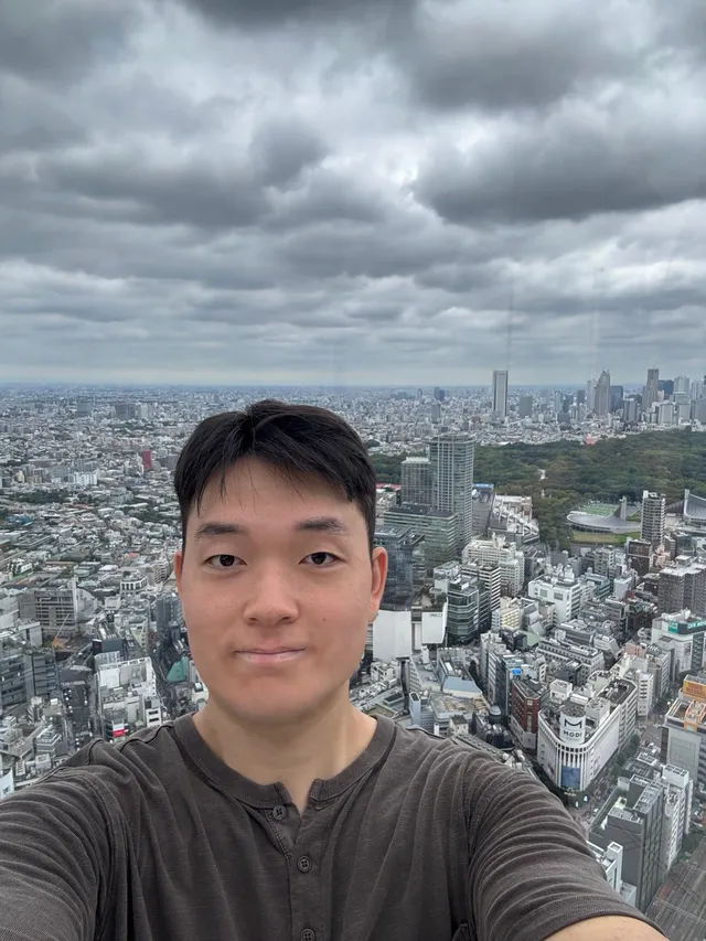 Doheon Kim taking a selfie at Shibuya Sky, with a wooded park and a stadium among the buildings behind him