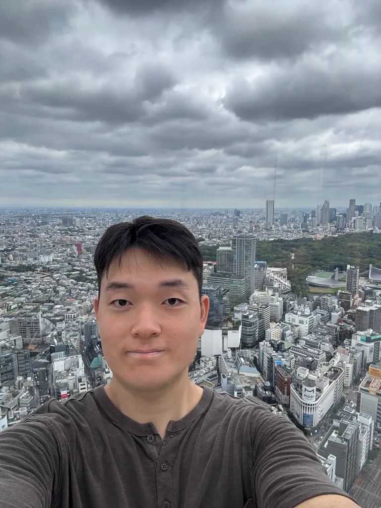 Doheon Kim taking a selfie at Shibuya Sky, with a wooded park and a stadium among the buildings behind him