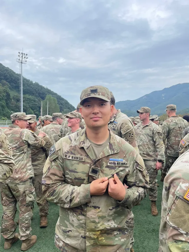 Doheon Kim holding the Expert Infantryman Badge pinned to his uniform, with other soldiers on the field behind him