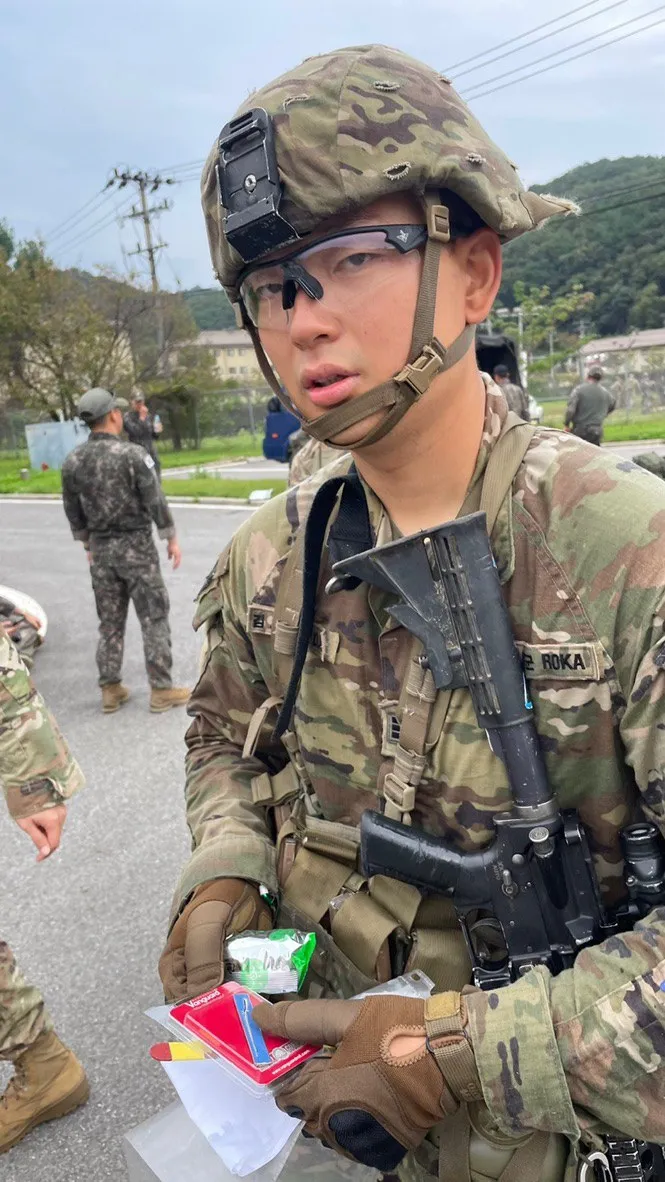 Doheon Kim in helmet, eye protection and gloves, carrying a rifle and holding snacks during testing