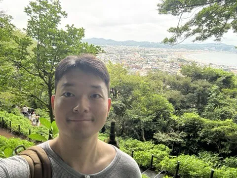 Doheon Kim taking a selfie on a wooded hillside, with a seaside town and bay below