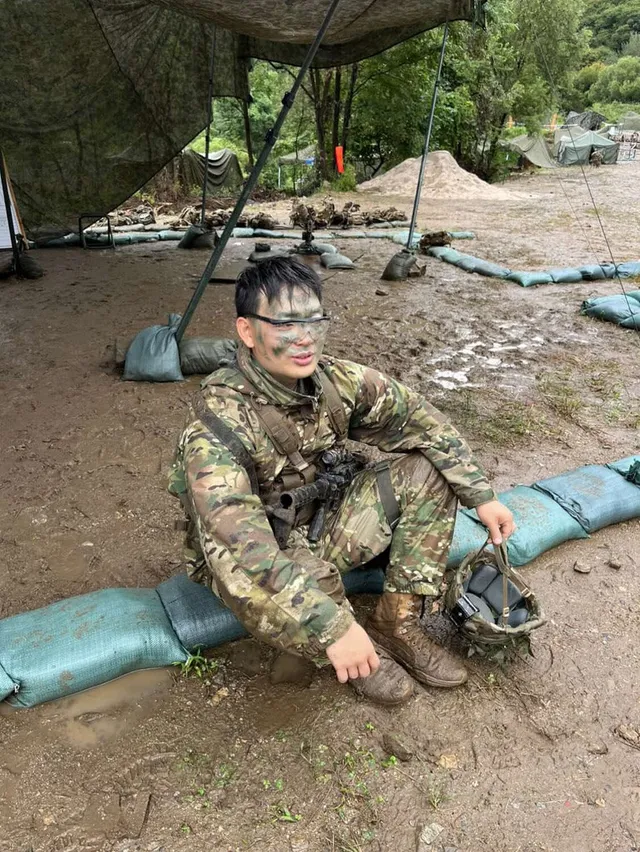 Doheon Kim in face paint, sitting on sandbags in the mud under camouflage netting with his helmet beside him