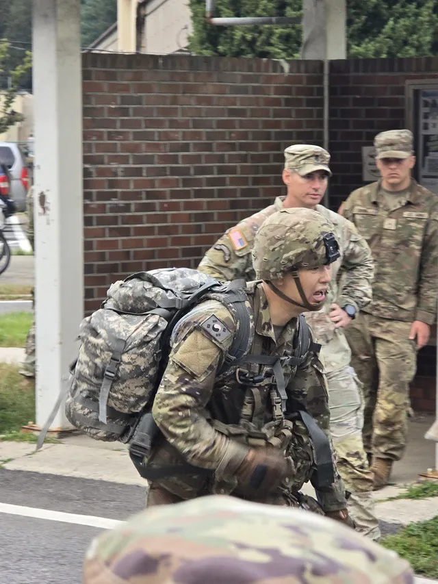 Doheon Kim bent forward under a full rucksack at the end of the ruck march, with two soldiers watching behind him
